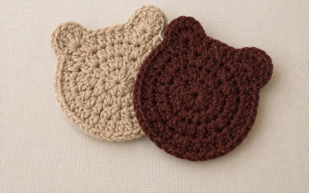 Two handmade bear-shaped crochet coasters in cream and chocolate brown on a neutral fabric background.
